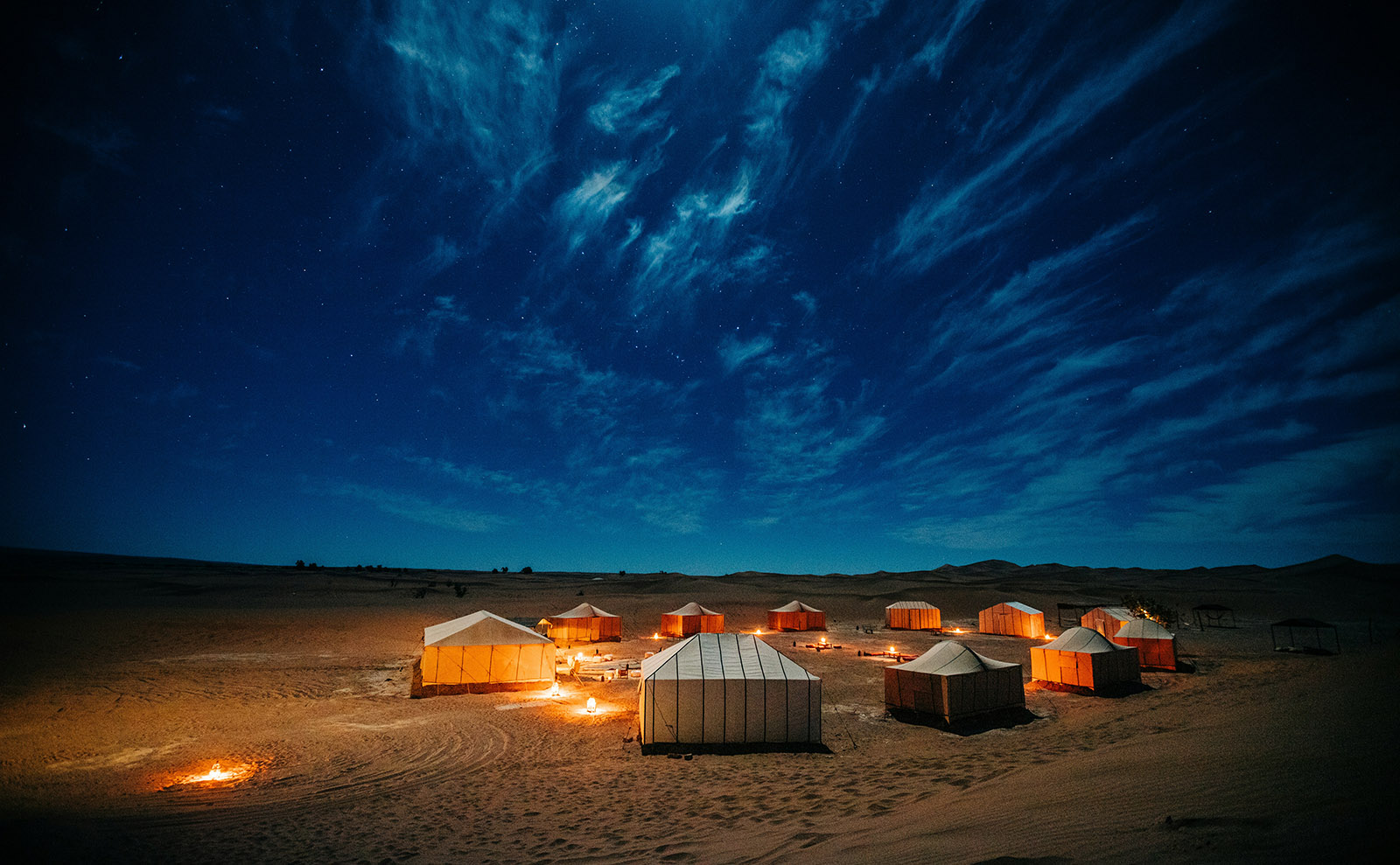 nighttime in the desert under a blue-black sky with golden sand and a circle of tents lit by torchlight