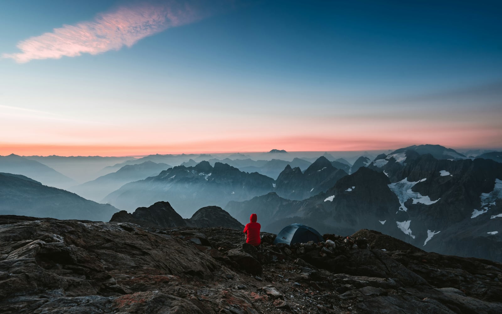 a man in a red jacket sits on gray rocks overlooking dozens of black rocky mountain tops