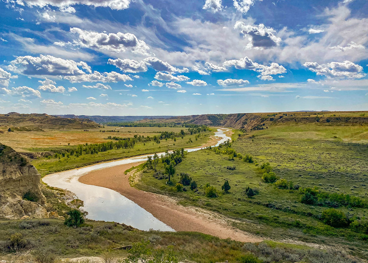 bright green grassy plains with a brown river running through the middle with blue sky and whispy white clouds overhead